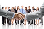 Two people wearing light colored suits have a firm handshake while a team of others stand in the background looking forward