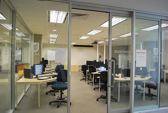 Rows of empty computer desks for team members are visible through an open doorway to an office behind a glass wall