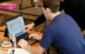 A person leans over a laptop on the desk while a co-worker points at the screen
