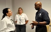 Two people with lanyards have a discussion in front of a doorway while another person stands behind them against the wall