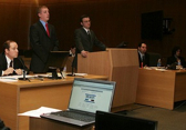A laptop sits open on a desk while multiple suited laywers are visible in the background of a courtroom