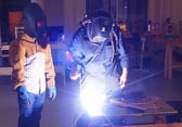A welder works on a piece of metal while an apprentice stands by to watch