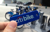 A person holds a blue tag between their fingers with the words citibike written on it with bicycles visible in the background