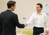 Two business people shake hands, one wearing a jacket and the other without