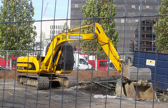 An excavator works in an open area, closed off by a fence