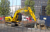An excavator works in an open area, closed off by a fence